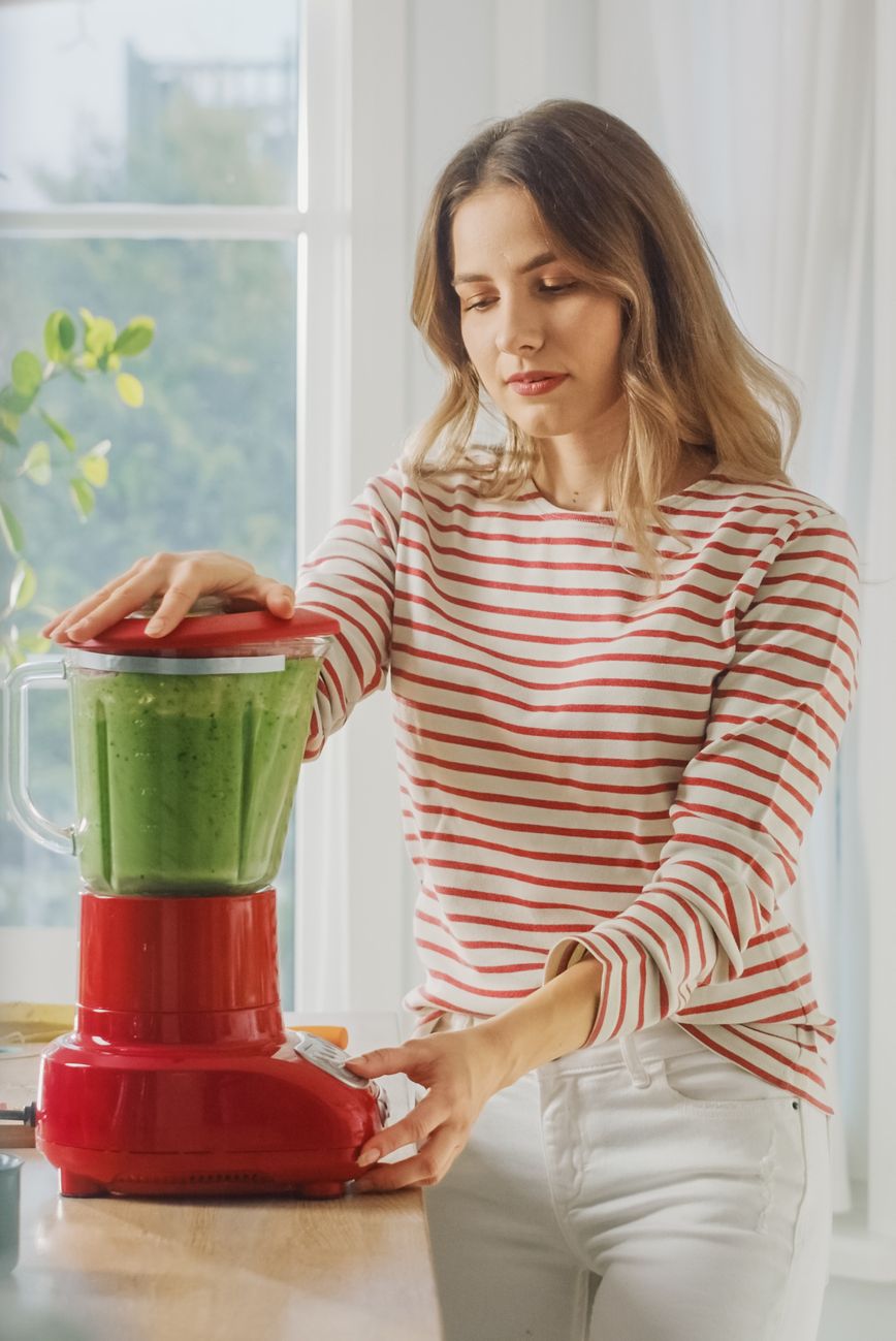 beautiful young female preparing a healthy green smoothie in a blender authentic stylish kitchen with healthy vegetables natural clean products from organic farming used to make drinks
