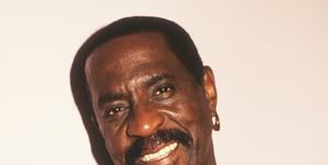 ike turner smiles at the camera, he wears a black shirt, a gold chain necklace and a gold hoop earring