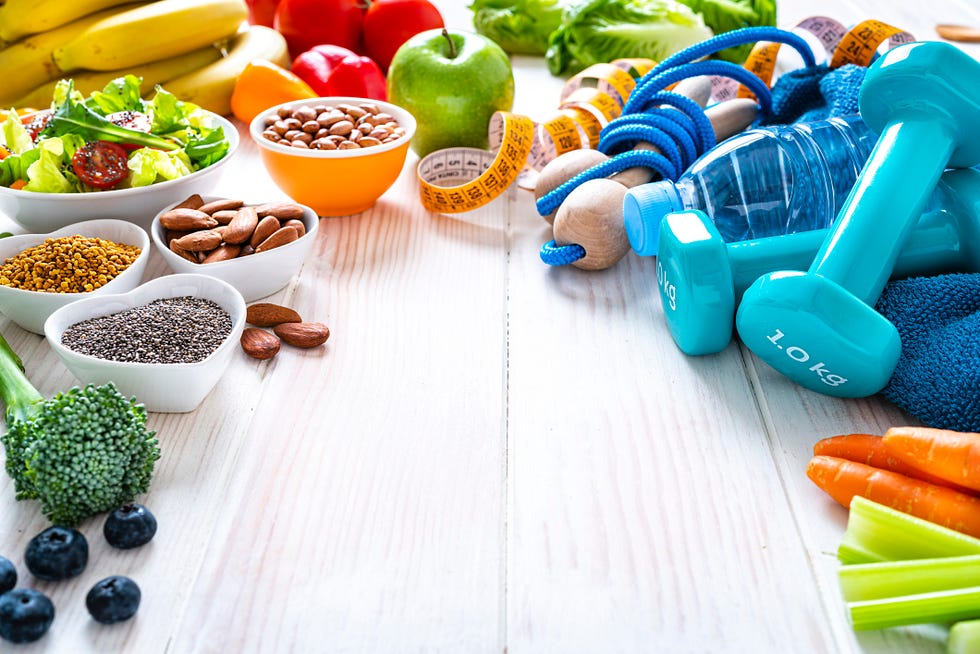 healthy eating and exercising concepts exercising items and healthy food shot on white table the composition arranged around the border of the frame leaving useful copy space and includes dumbbells, blue towel, jump rope, tape measure, a water bottle and some fruits, vegetables and nuts like broccoli, celery, almonds, carrots, blueberries, a green apple, chia seeds, bananas, pinto beans, tomatoes and pistachio a salad bowl with lettuce, cherry tomatoes, cucumber, and carrot sticks is at the left high resolution 42mp studio digital capture taken with sony a7rii and zeiss batis 40mm f20 cf lens