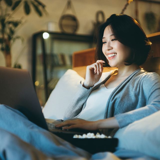 young asian woman lying on the bed enjoying the weekend, watching movie on laptop and eating popcorn at home in the evening
