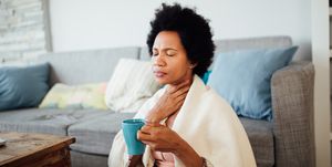 african american woman, fallen ill is staying at home wrapped in a blanket socially distancing and quarantining herself, feeling her throat hurt and being sore, having a cup of hot tea
