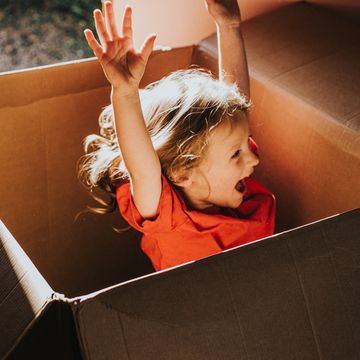 cute and joyful little girl excitedly leaps inside a huge cardboard box in sunlight she throws her hands up in excitement background image with child being in soft focus, and box provides a space for copy