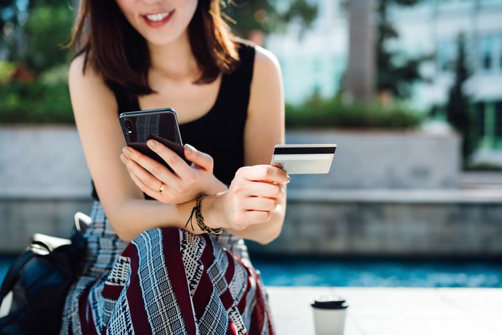 beautiful smiling young asian woman sitting in an urban park, enjoying coffee, shopping online with smartphone and making mobile payment with credit card on hand online shopping and mobile payment concept