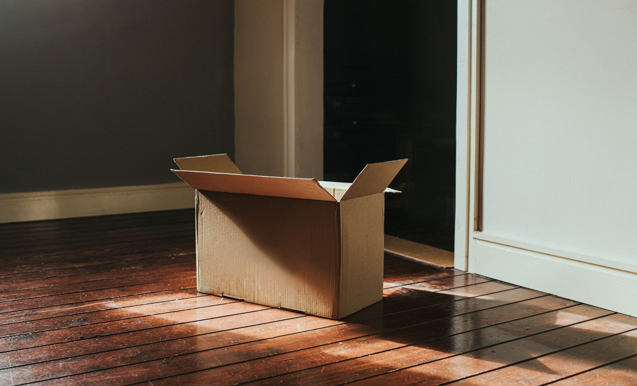 a single, plain, cardboard box sits on a dark wooden floor in a domestic room sun shines through the window creating flares and shadows room is empty and sparse wall provides a space for copy