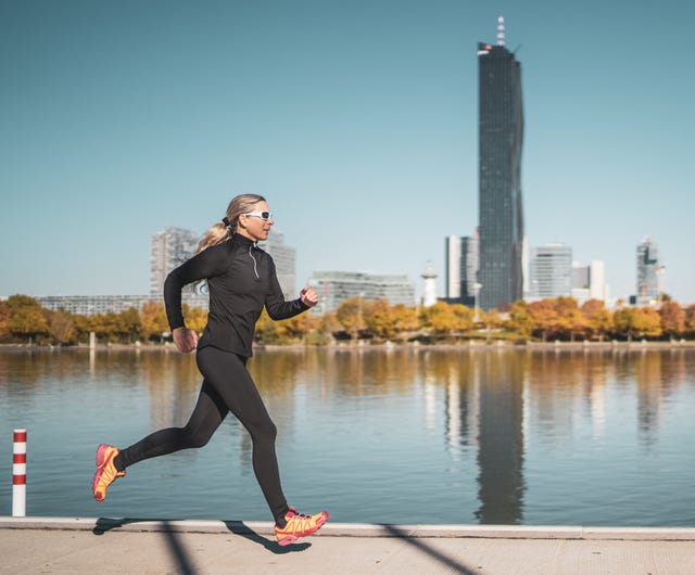 side view female mature adult athlete jogging alone along riverside in city on sunny day in autumn with urban skyline in background