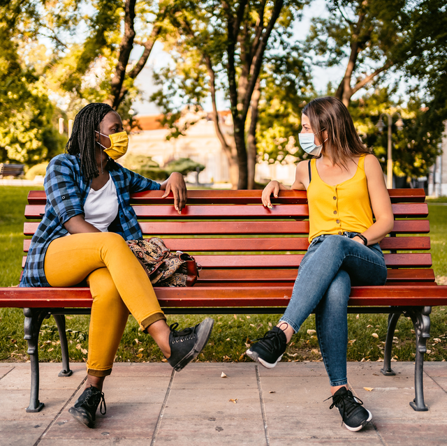 two friends in face masks sitting on opposite sides of bright red bench in park