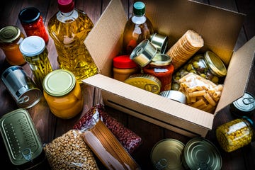 high angle view of a cardboard box filled with multicolored non perishable canned goods, conserves, sauces and oils shot on wooden table. the composition includes cooking oil bottle, pasta, crackers, preserves and tins. high resolution 42mp studio digital capture taken with sony a7rii and zeiss batis 40mm f2.0 cf lens