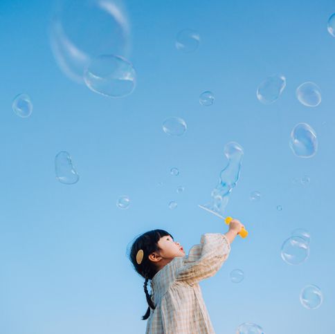 adorable little asian girl holding a bubble wand having fun playing with soap bubbles outdoors in nature park against beautiful blue sky on a lovely day