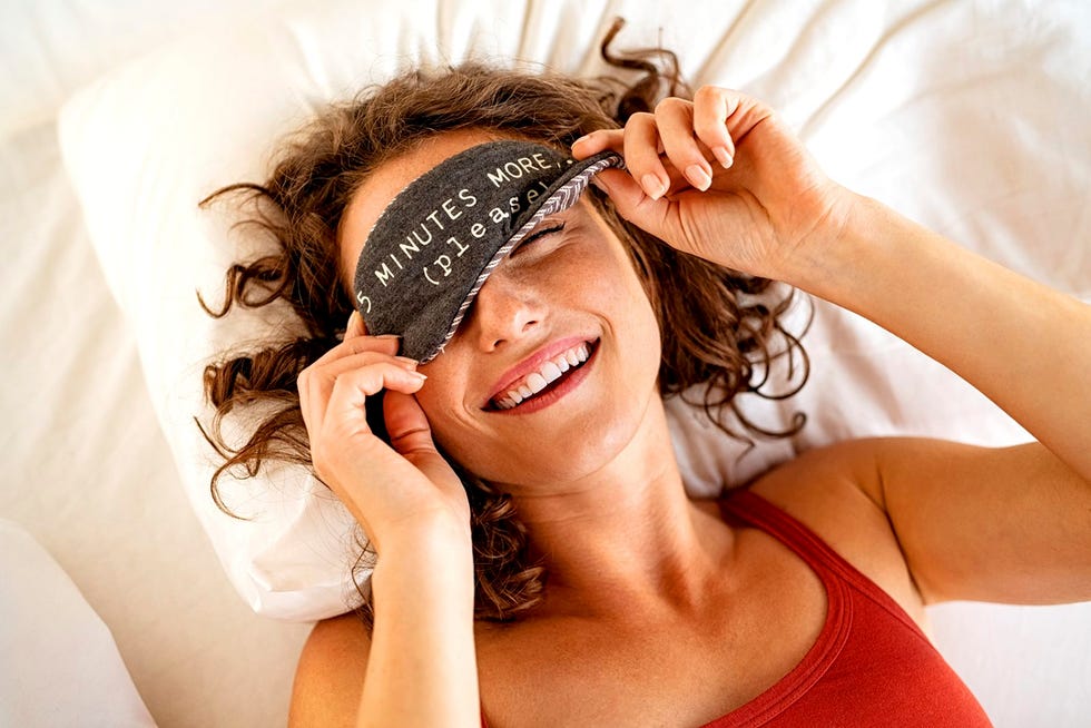 top view of tired young woman sleeping on bed close up face of beautiful sleepy girl wearing funny eye mask while peeping with one eye high angle view of woman covering her eyes with sleeping mask with five minutes more text