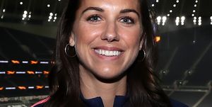 alex morgan smiles at the camera while standing inside a stadium, she wears silver hoop earrings and a navy jacket