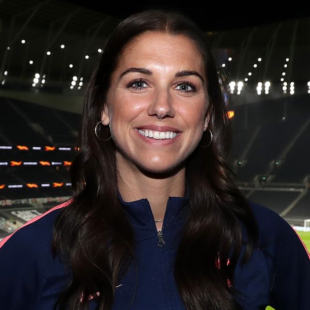 alex morgan smiles at the camera while standing inside a stadium, she wears silver hoop earrings and a navy jacket