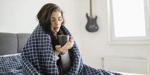 een zieke vrouw zit onder een dekentje en houdt een kop thee vast