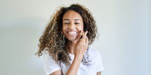 cheerful young woman applying facial cream against white wall