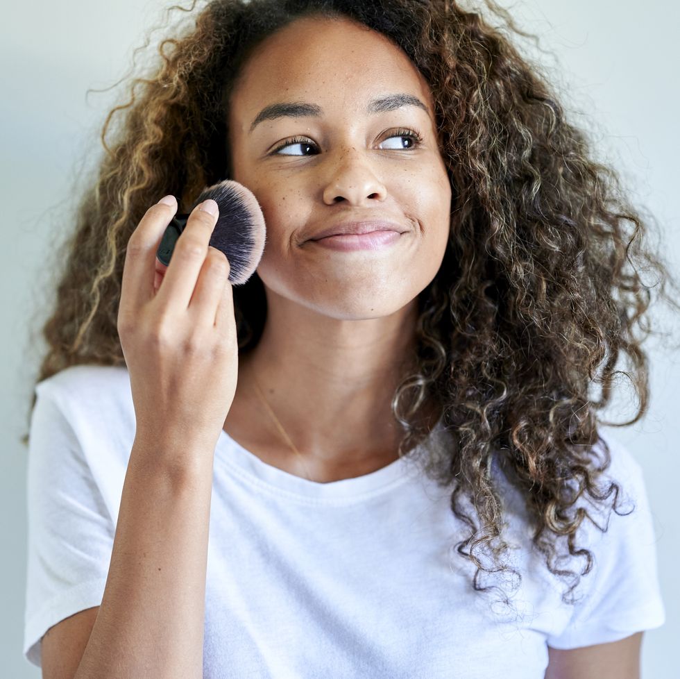 smiling young woman applying face powder with make up brush against white wall