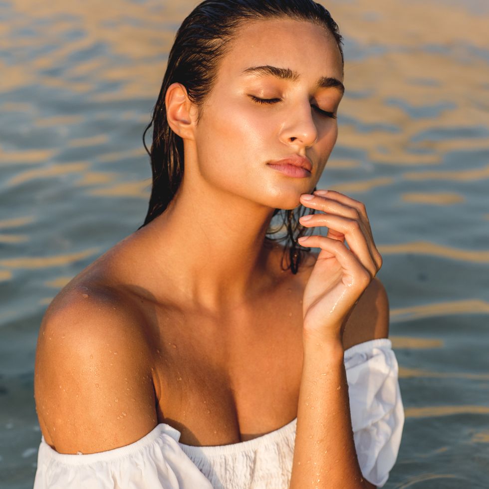 portrait of exotic beautiful woman standing in the ocean water wearing a wet white sleeveless top during sunset