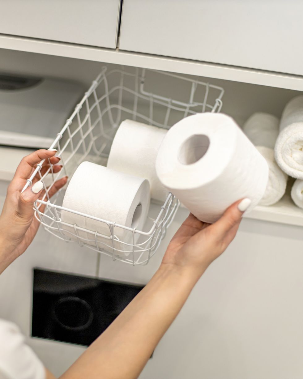 young woman with a smile is organizing and placing mesh basket with stack of toilet paper rolls in bathroom cupboard near rolled up hand towels happy housewife is tidying up toiletries in bathroom
