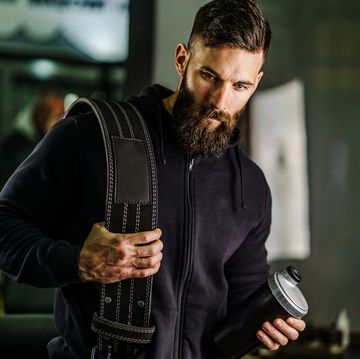front view portrait of young caucasian man athlete in black hoodie male standing in the gym holding protein supplement shaker and belt supplementation in training black hair and beard copy space