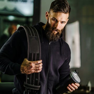 front view portrait of young caucasian man athlete in black hoodie male standing in the gym holding protein supplement shaker and belt supplementation in training black hair and beard copy space