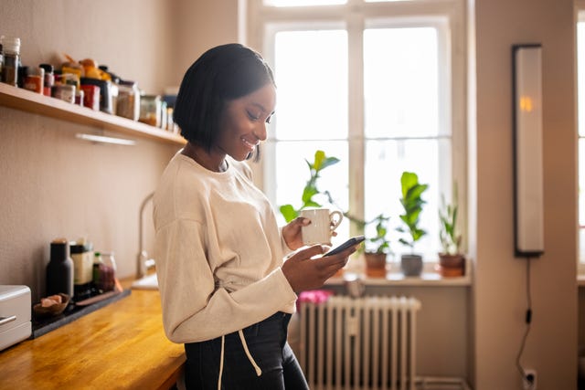 woman standing in the kitchen holding a cup of coffee and texting on her cell phone woman using a mobile phone at home