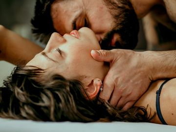 side view photo of a beautiful couple in bed, being gentle and sensual handsome guy with a beard on top of the beautiful brunette is kissing her neck while her eyes are closed making love concept