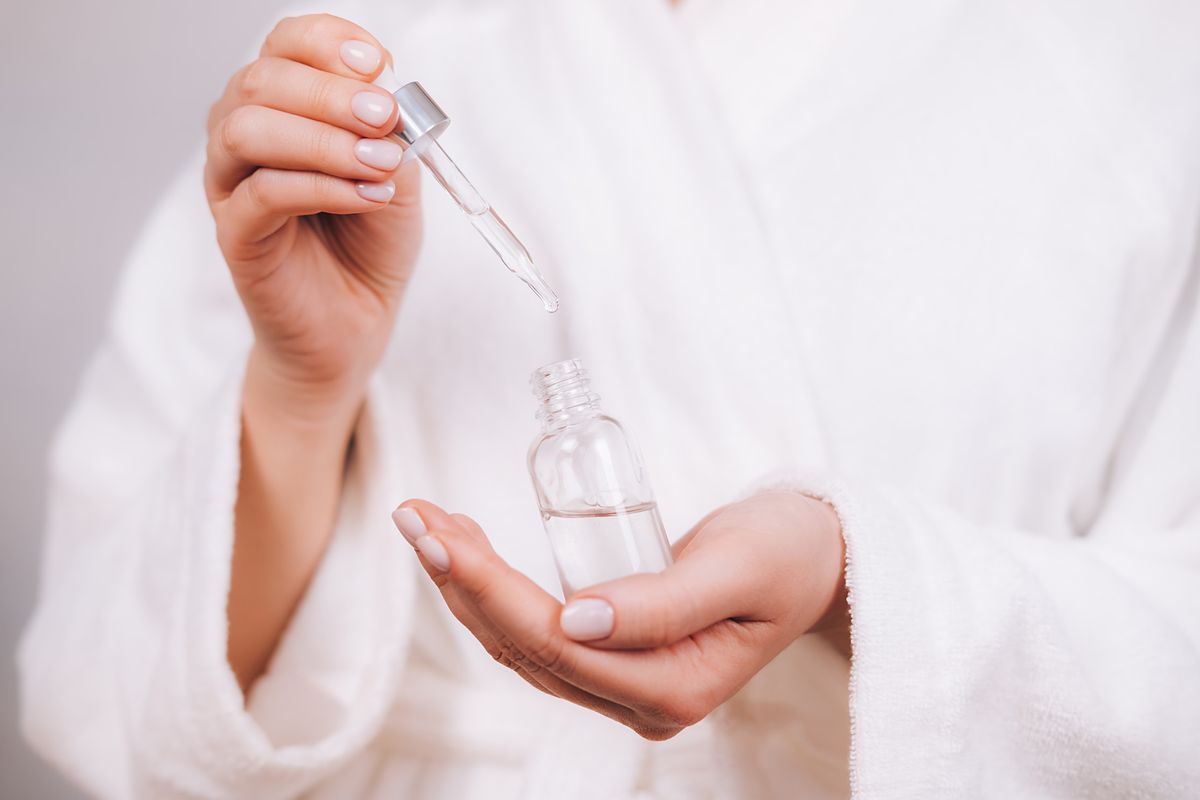 woman in white bathrobe with nude pink manicure holds pipette and transparent glass bottle with natural essential oil or organic serum she ready to start skin care procedures concept of home body care and healthy lifestyle close up front view