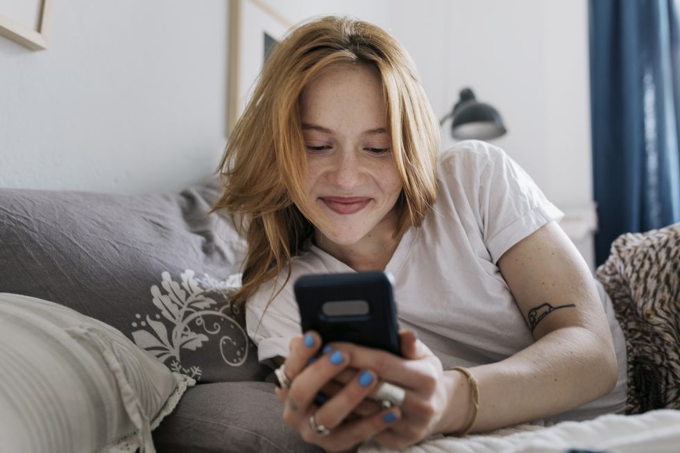 a woman smiling while lying on her bed at home and messaging on an online dating app using her smartphone