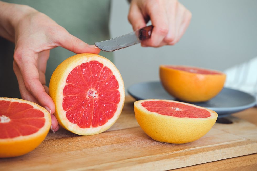 a girl or woman is holding a knife, cutting a red ripe grapefruit in half, on a cutting board, against the background of a wooden kitchen table there is a manual juicer nearby making juice for breakfast the concept of vegetarian, vegan and raw food