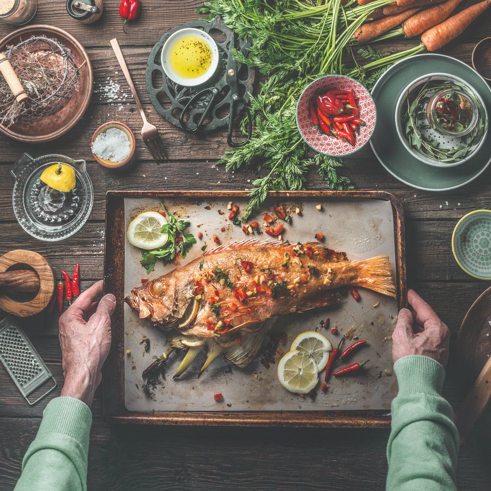 mans hands holding roasted whole redfish stuffed with fennel and lemon on baking tray on rustic table with fresh ingredients and kitchen utensils top view healthy food concept home cuisine