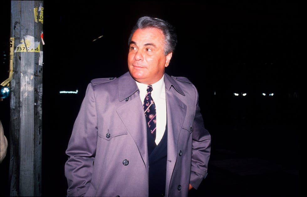john gotti looks over his shoulder while walking outside at night, he wears a gray coat over a suit and tie