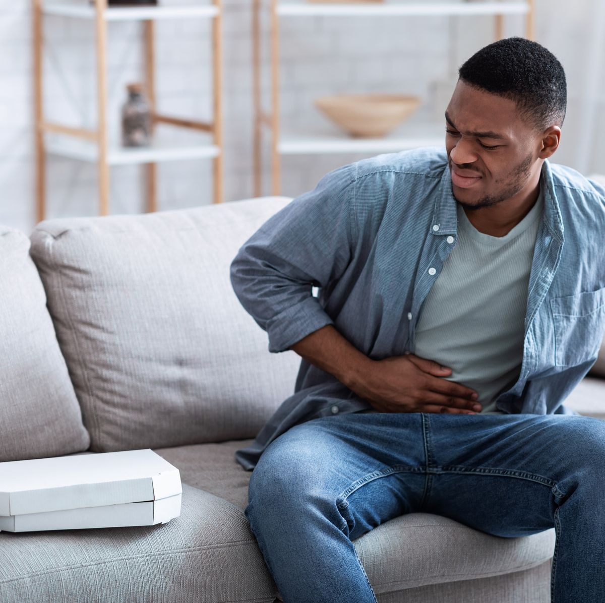 african american guy having stomachache after eating pizza touching aching stomach suffering from pain sitting on sofa at home food poisoning, gastritis, abdominal inflammation and appendicitis