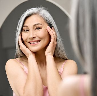 happy middle 50 years aged asian woman with gray hair looking at mirror reflection examining touching face enjoying antiaging beauty treatments beauty hydrate skin care wrinkle prevention concept