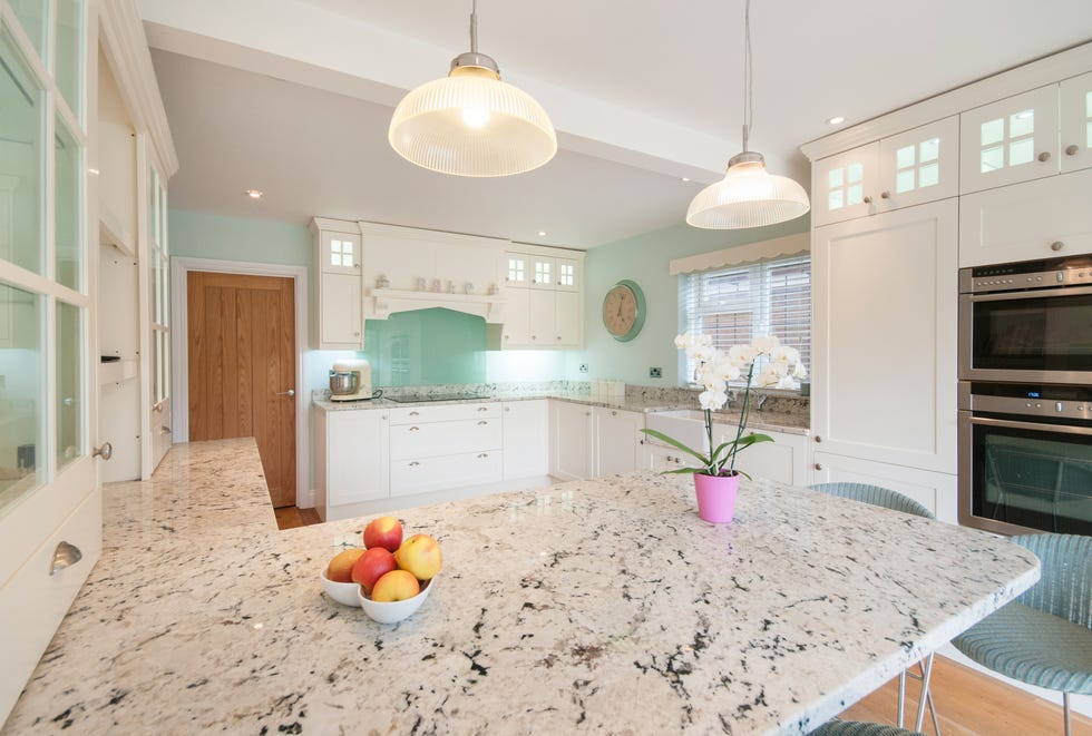 a general interior view of a white fitted shaker style kitchen with island, wicker bar stools, glass shade ceiling lights, potted orchid and fruit bowl