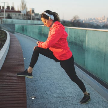 Woman in headphones warming up and stretching before morning run on city quay. Active and healthy life young pretty girl in red jersey training outdoors in morning city urban view wellbeing and vitality, power in life workout outside in town beautiful sunrise light special running exercises millennial generation