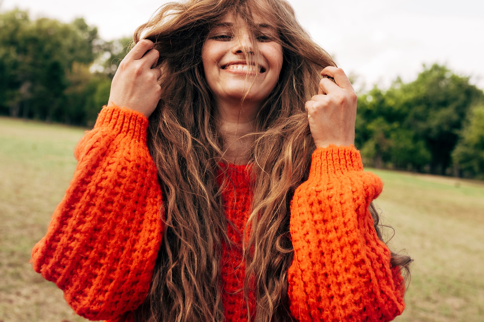 positive young woman playing with her long red hair wearing an orange knitted sweater posing on the nature background pretty female playing with her hair outdoors in the park