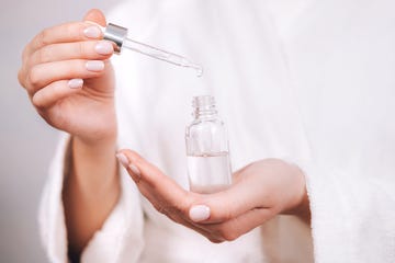 woman in white bathrobe with nude pink manicure holds pipette and transparent glass bottle with natural essential oil or organic serum she ready to start skin care procedures concept of home body care and healthy lifestyle close up front view