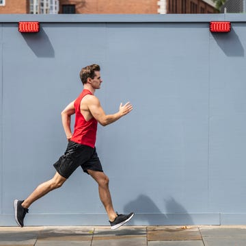 Fit young man running in urban neighbourhood full length side view of caucasian man in red singlet, black shorts and shoes, striding along sidewalk during summertime weekend workout.