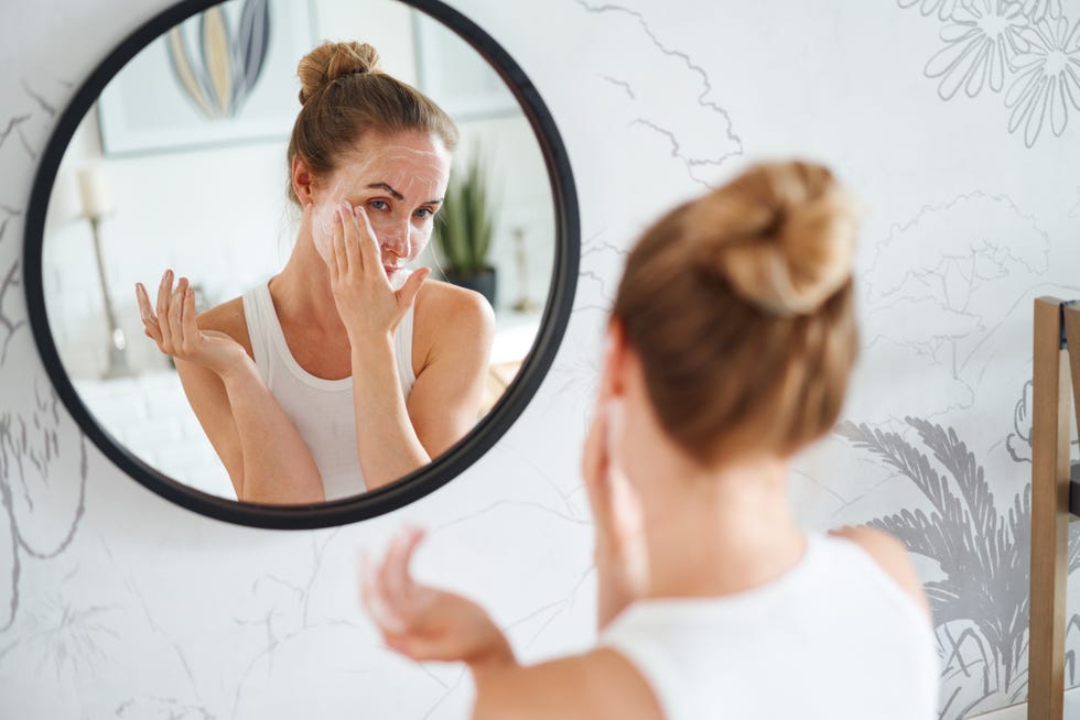 Young woman reflecting in mirror, applying cleansing cosmetic mask on face during skincare routine in bathroom