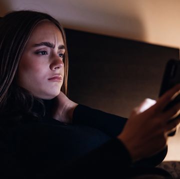 worried and disappointed looking young woman lying on her bed in illuminated bedroom at night reading bad news in her e mails, chat messages or social media posts on her mobile phone ambient bedroom night lighting millenial generation modern technology lifestyle