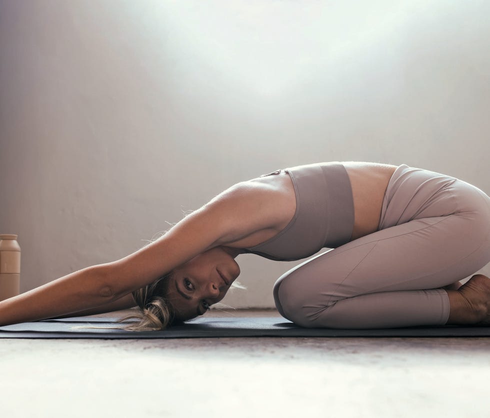 a close up of a young blonde caucasian woman stretching on her yoga mat holding a child's pose with an eco friendly water bottle barefoot