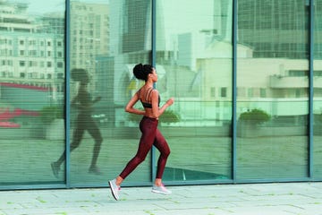 young sportswoman in stylish tracksuit runs past modern building with glass facade reflecting large city street side view