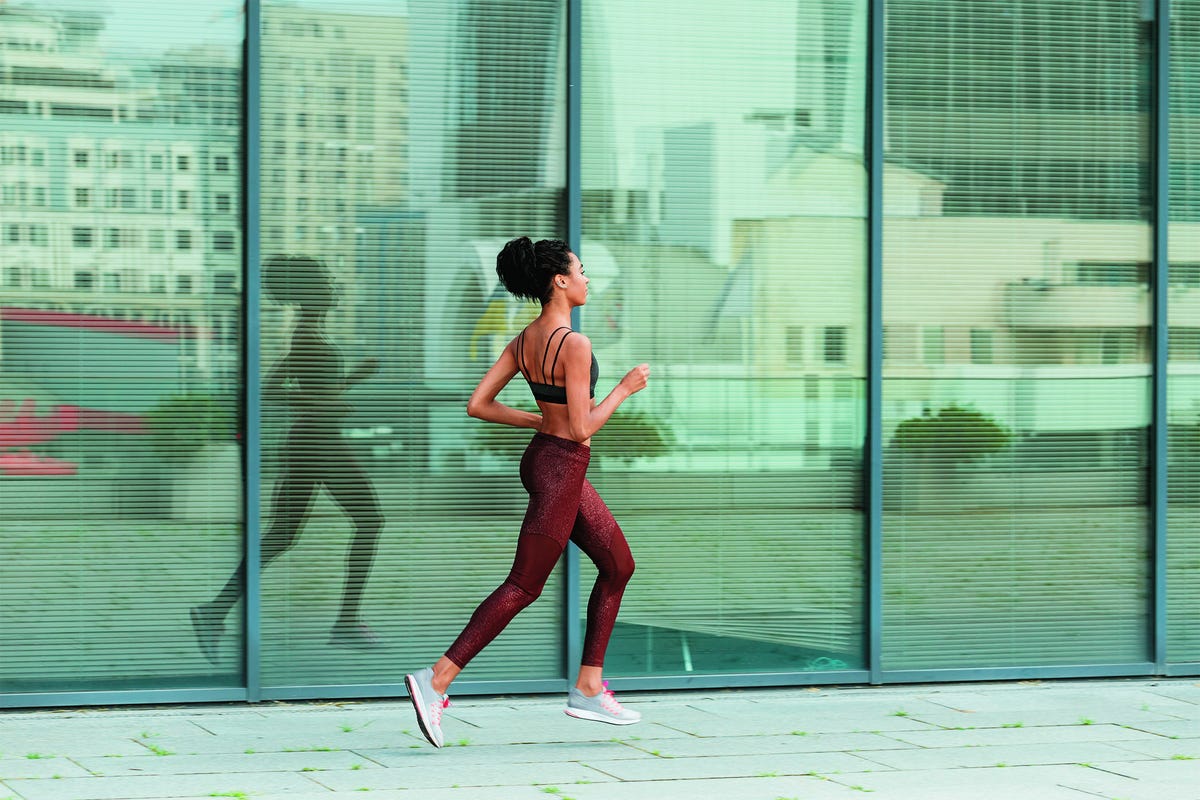 young sportswoman in stylish tracksuit runs past modern building with glass facade reflecting large city street side view