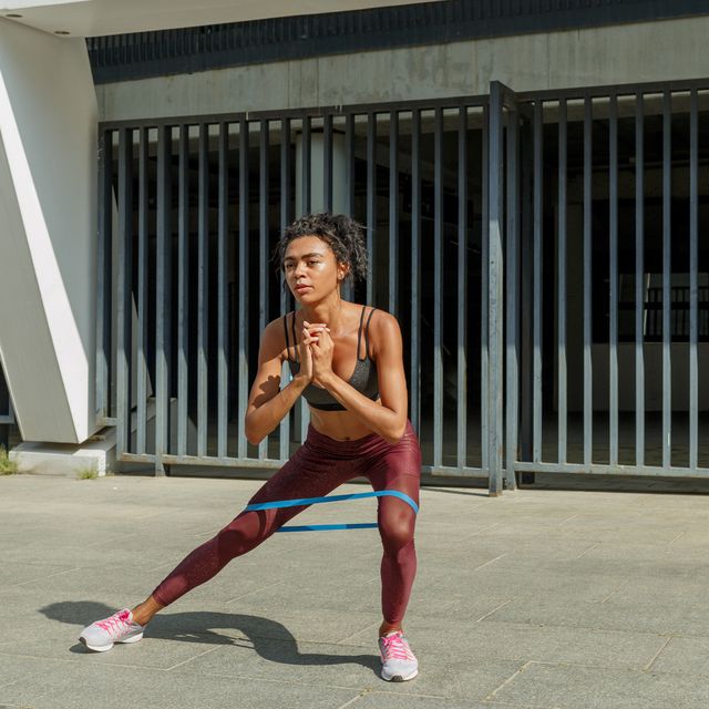 attractive young  woman in stylish tracksuit does side squats with elastic band and keeps hands on waist near modern building on sunny city street