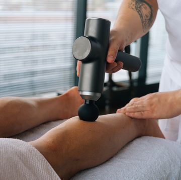 close up of unrecognizable professional male masseur massaging leg calf muscles using massage gun percussion tool of muscular athlete man, on spa treatment lying on back in massage table
