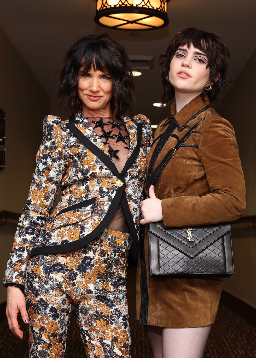 juliette lewis, in a black, white, and gold floral suit, and sophie thatcher, wearing a brown leather dress, pose together in a hallway