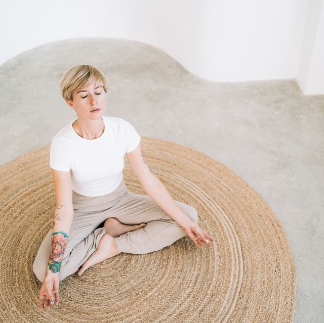 beautiful authentic woman with tattoos and short blond hair is meditating sitting in lotus position on wicker carpet in minimalist home interior she is wearing a light colored casual clothing concept of relaxation exercises