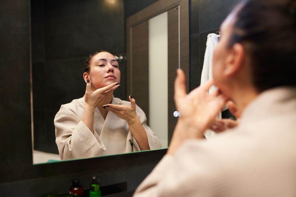 woman in cotton bathrobe applying cream and massaging chin while looking at mirror during beauty routine in bathroom at home