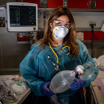 a female doctor in scrubs, goggles, and a mask looks directly into the camera in a hospital room