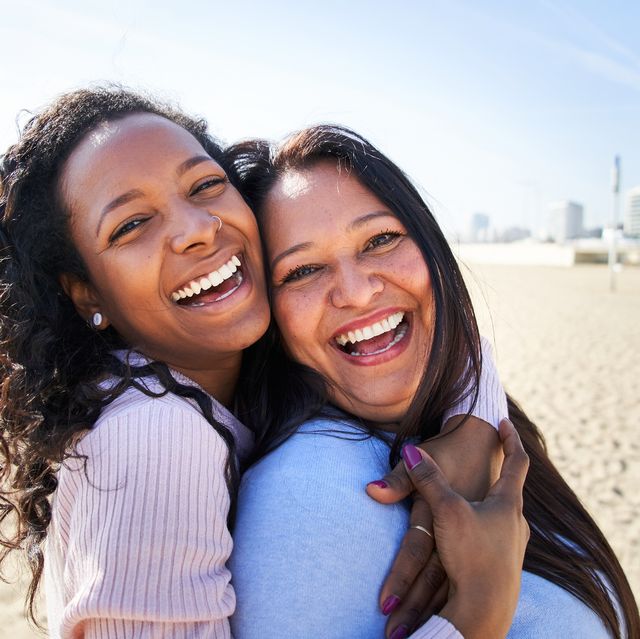 close up photography of multiracial family, looking and smiling at camera, daughter hugging her mother, standing on the beach, woth big smile