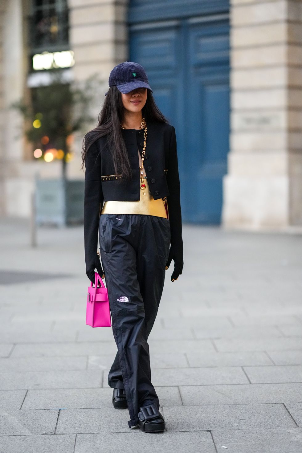 paris, france march 06 a guest wears a navy blue ribbed velvet cap from new era, a gold and rhinestones long pendant necklace from schiaparelli, a gold shiny leather with embroidered gold jewel corset from schiaparelli, a black cropped jacket from schiaparelli, black gloves from schiaparelli, a pink shiny leather handbag, black large pants from the north face, black shiny leather with buckle on the toe cap shoes, outside schiaparelli, during paris fashion week womenswear fw 2022 2023, on march 06, 2022 in paris, france photo by edward berthelotgetty images