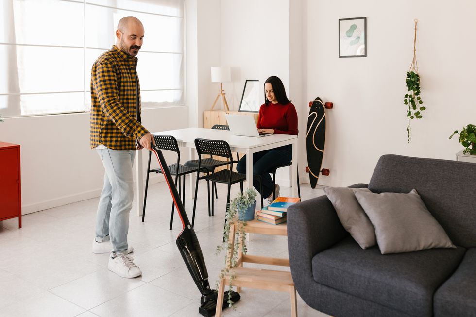 man using a vacuum cleaner to cleaning his living room floor while his girlfriend working on a laptop home office, cleanliness and hygiene conceptman using a vacuum cleaner to cleaning his living room floor while his girlfriend working on a laptop housework, domestic life and home office concept